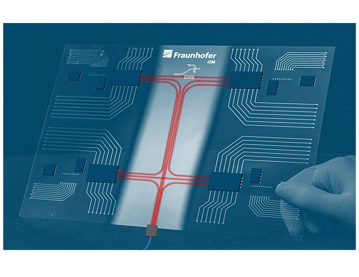 Eine Hand hält eine transparente Leiterplatte mit aufgedruckten roten Licht- bzw. Datenwegen, die sich in der Mitte kreuzen. Oben ist das Fraunhofer‑Logo sichtbar, das auf die wissenschaftliche Herkunft der Technologie hinweist.
