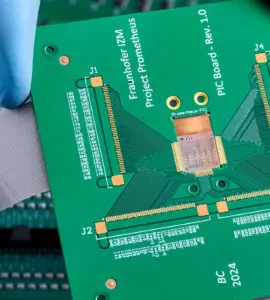 A gloved hand holds a green printed circuit board (PCB) above other electronic circuit boards with connectors and ribbon cables, set against a dramatically cloudy background.