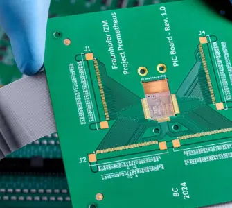 A gloved hand holds a green printed circuit board (PCB) above other electronic circuit boards with connectors and ribbon cables, set against a dramatically cloudy background.