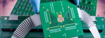 A gloved hand holds a green printed circuit board (PCB) above other electronic circuit boards with connectors and ribbon cables, set against a dramatically cloudy background.