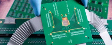 A gloved hand holds a green printed circuit board (PCB) above other electronic circuit boards with connectors and ribbon cables, set against a dramatically cloudy background.