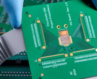A gloved hand holds a green printed circuit board (PCB) above other electronic circuit boards with connectors and ribbon cables, set against a dramatically cloudy background.