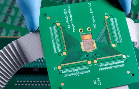 A gloved hand holds a green printed circuit board (PCB) above other electronic circuit boards with connectors and ribbon cables, set against a dramatically cloudy background.
