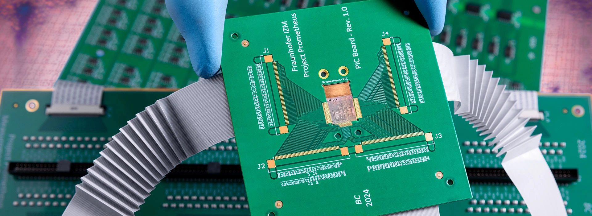 A gloved hand holds a green printed circuit board (PCB) above other electronic circuit boards with connectors and ribbon cables, set against a dramatically cloudy background.