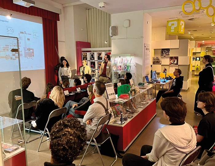 Gruppe von Menschen sitzt in einem Geschäft mit gelber Deckendekoration und roten Regalen und hört einer Präsentation zu.
